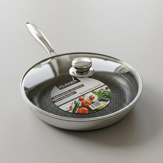 Studio shot of a stainless steel honeycomb frypan with a glass lid and branded insert card resting on a marble kitchen countertop.